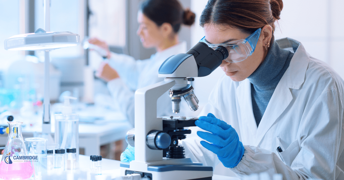 a female chemist looks into a microscope in front of an array of scientific testing equipment in a lab