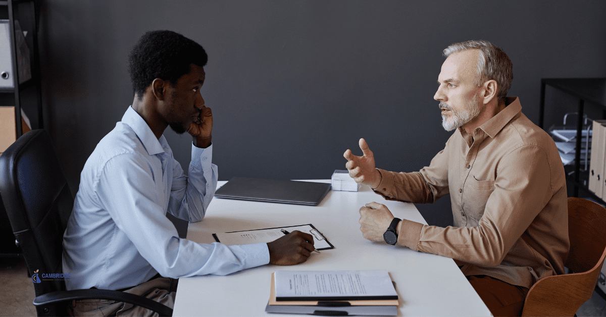 Two men sitting at a desk in an office. The young man on the left is interviewing the older man on the right.