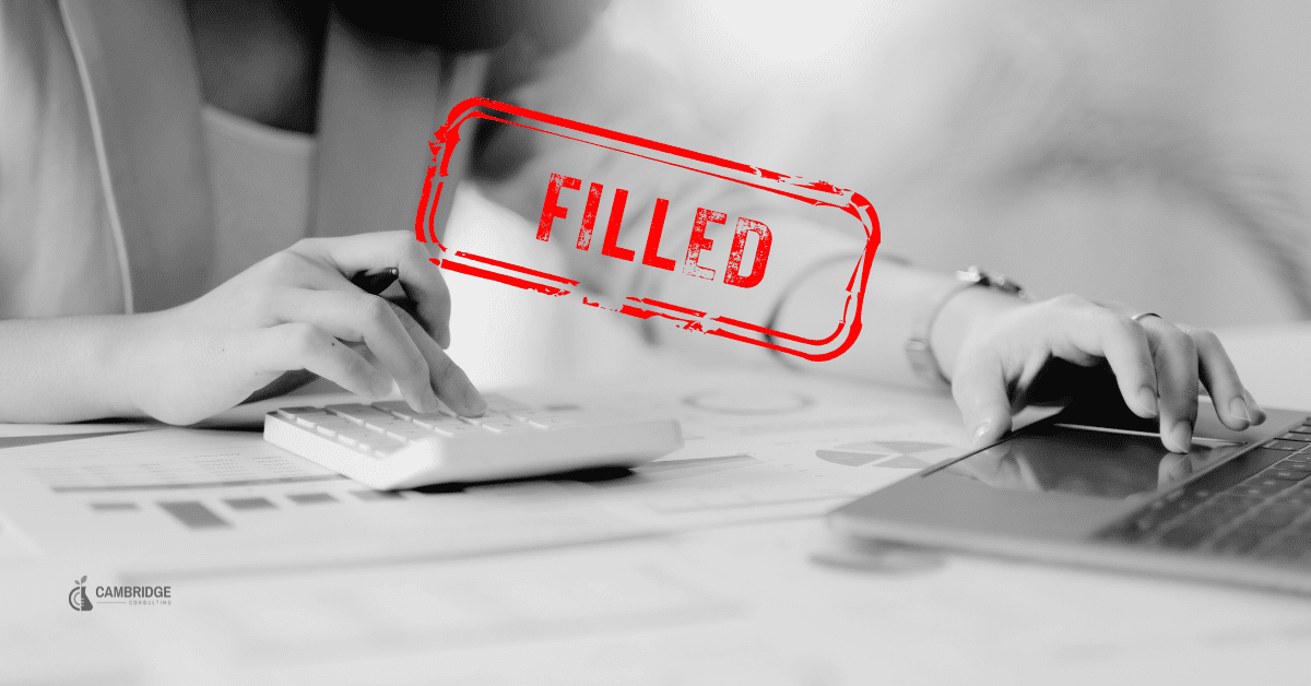 A black and white image of a woman typing numbers into a calculator and writing in a note pad at her desk with a red stamp saying "filled" across the image.