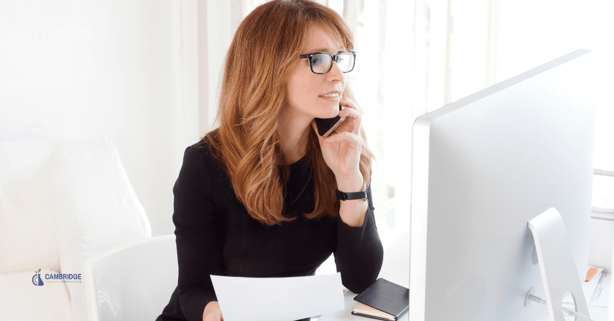 corporate woman sits in her office talking with a client on the phone as she holds a piece of paper and looks at her computer screen.