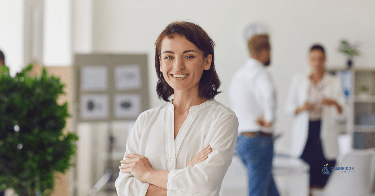 Corporate woman smiling with her hands crossed over her chest with two colleagues chatting in the background.