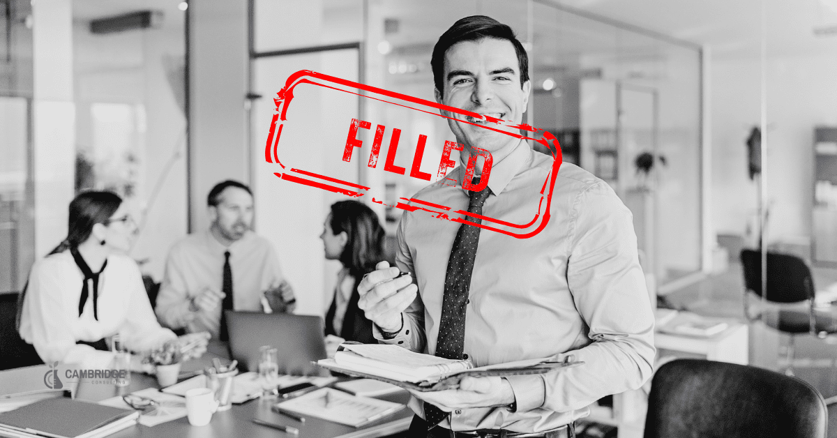 a black and white image of a corporate man smiling in front of his work colleagues with a red stamp overlay that says "FILLED"
